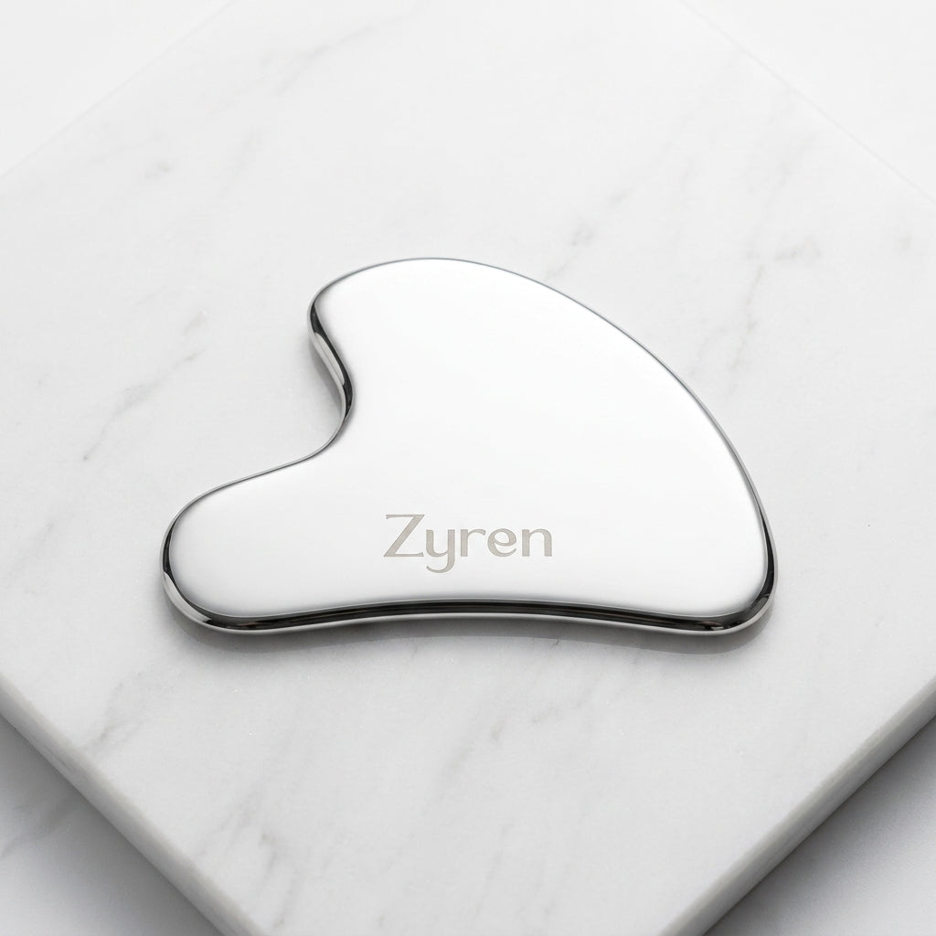 Zyren Gua Sha, a polished metal facial tool engraved with the Zyren logo, lying on white marble